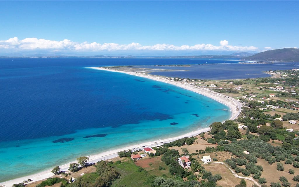 Vista aerea di Agios Ioannis, Lefkada, con spiaggia dorata e mare turchese in una mattina estiva prima dell’arrivo dei kite surfer. Mulini a vento e vegetazione mediterranea sullo sfondo.