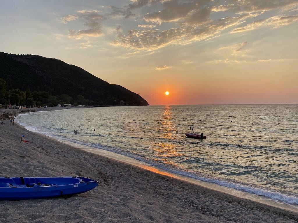 Tramonto estivo sulla spiaggia di Agios Ioannis a Lefkada, con il cielo infuocato che si riflette sul mare calmo e sulla sabbia dorata.