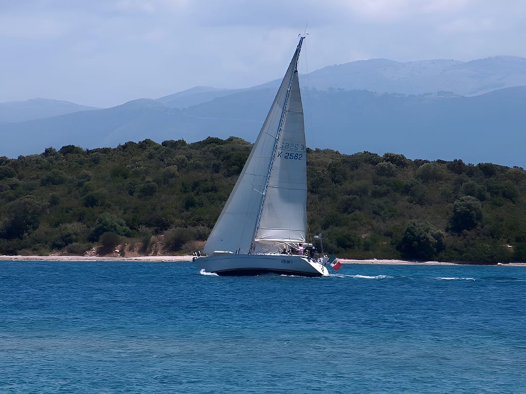 Una elegante barca a vela con bandiera italiana naviga sottocosta a Meganissi, tra acque cristalline e paesaggi incantevoli.