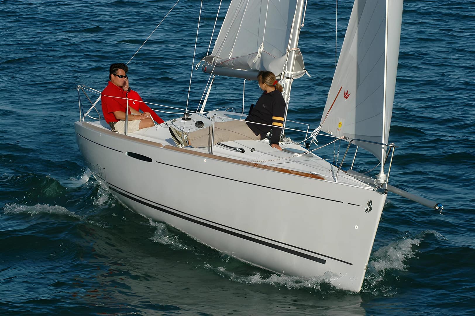 Un Beneteau First 21.7 solca il mare calmo con le vele completamente spiegate, tre persone a bordo immerse nell’esperienza della navigazione.