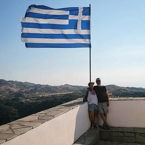 Due turisti sorridenti immortalati sotto la bandiera greca che sventola sopra di loro, un ricordo speciale della visita al punto panoramico di Profiti Ilias, con una vista panoramica sull'isola e sul mare circostante.