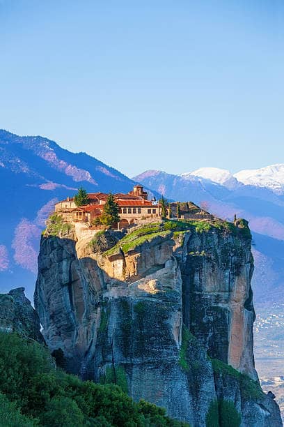 Veduta verticale del monastero di Meteora, avvolto nella luce dorata del mattino, con una maestosa catena montuosa sullo sfondo e un cielo terso che crea un suggestivo contrasto.