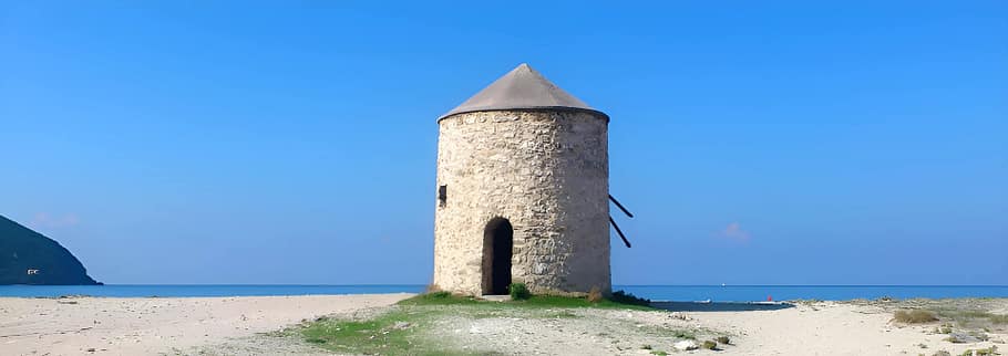 Uno dei mulini storici sulla spiaggia di Agios Ioannis, incorniciato dalla linea dell’orizzonte marino, la sabbia bianca e il promontorio che emerge sulla sinistra.